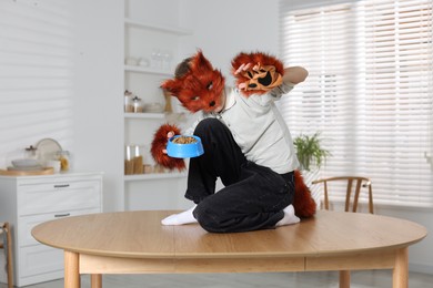 Photo of Quadrobics. Boy wearing fox mask, tail and gloves with feeding bowl on table indoors