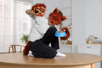 Photo of Quadrobics. Boy wearing fox mask, tail and gloves with feeding bowl on table indoors