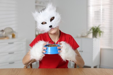 Photo of Quadrobics. Boy wearing cat mask and gloves with feeding bowl at table indoors