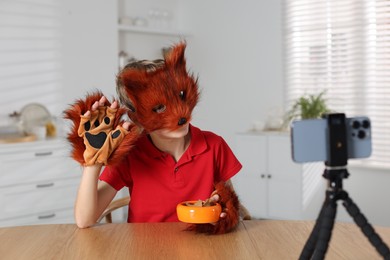 Quadrobics. Boy wearing fox mask and gloves with feeding bowl filming video at table indoors