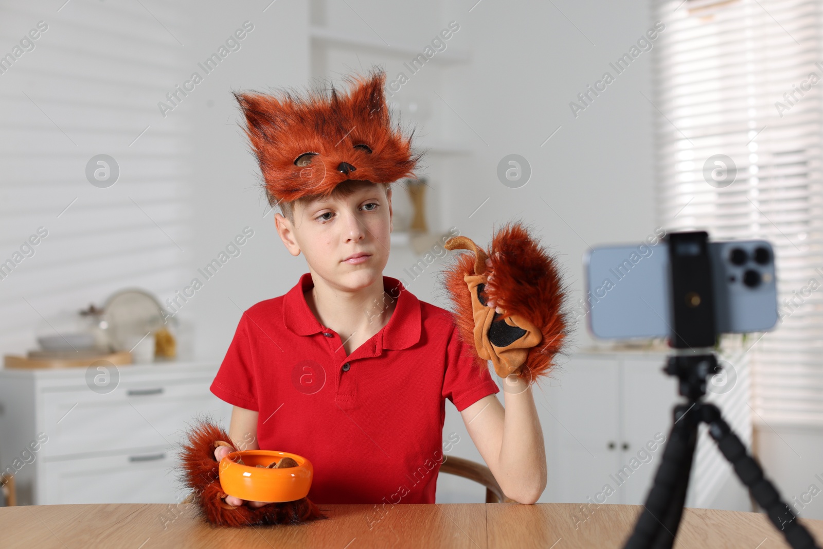 Photo of Quadrobics. Boy wearing fox mask and gloves with feeding bowl filming video at table indoors