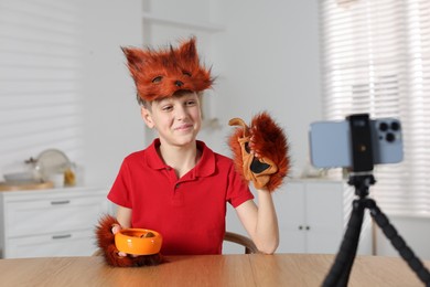 Photo of Quadrobics. Boy wearing fox mask and gloves with feeding bowl filming video at table indoors