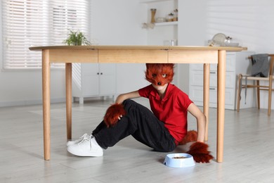 Photo of Quadrobics. Boy wearing fox mask, tail and gloves with feeding bowl under table indoors