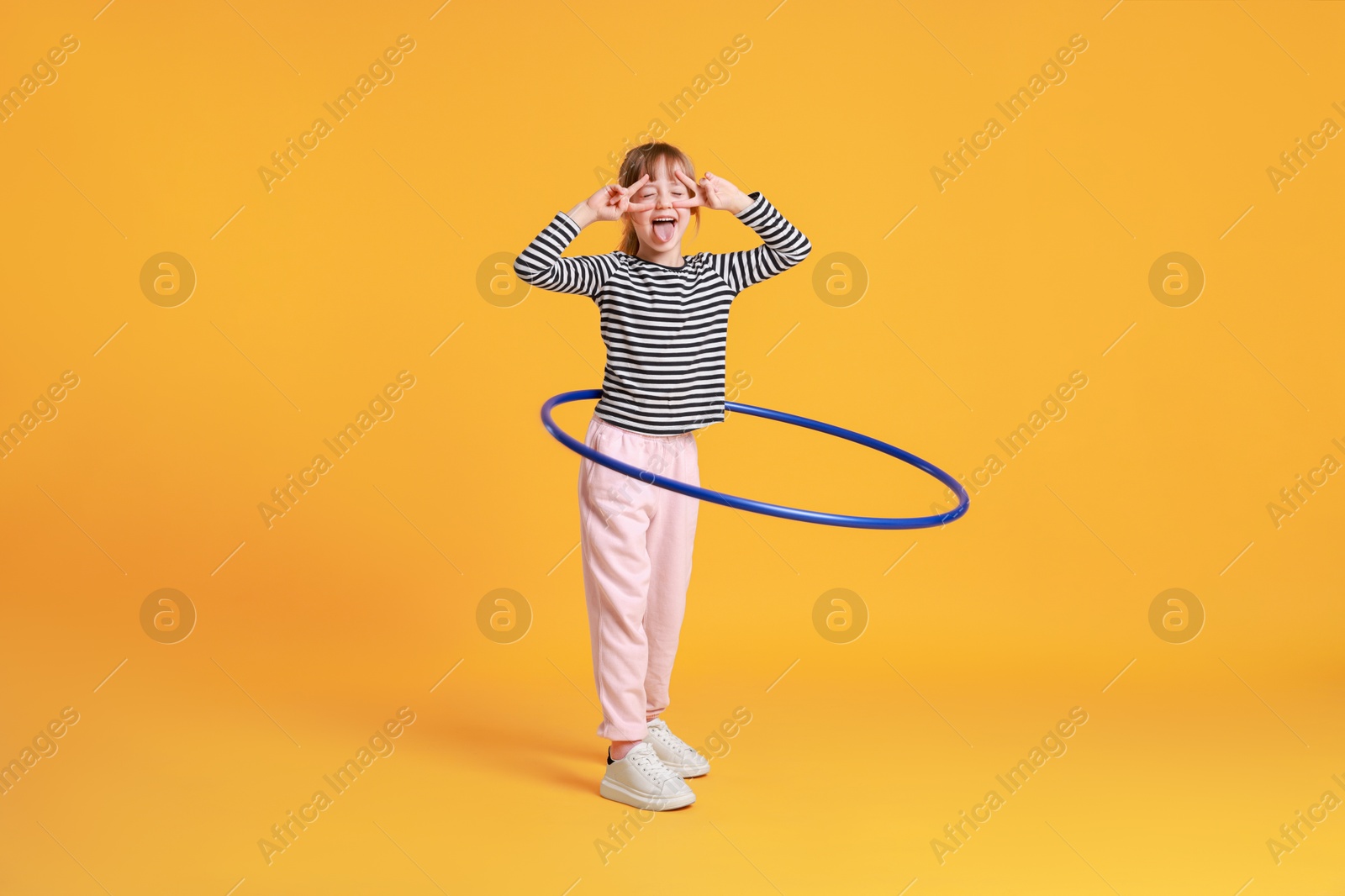 Photo of Cute little girl showing V-signs while exercising with hula hoop on orange background