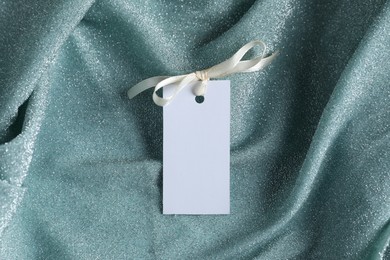 Photo of Blank tag with ribbon bow on teal fabric, top view