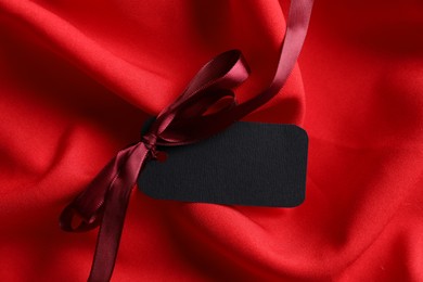 Photo of Blank tag with ribbon bow on red fabric, closeup