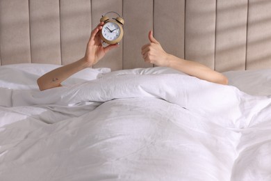 Photo of Woman with alarm clock showing thumbs up in bed at home, closeup. Good morning