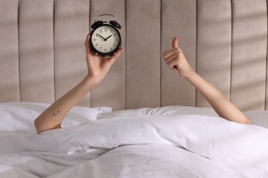 Photo of Woman with alarm clock showing thumbs up in bed at home, closeup. Good morning