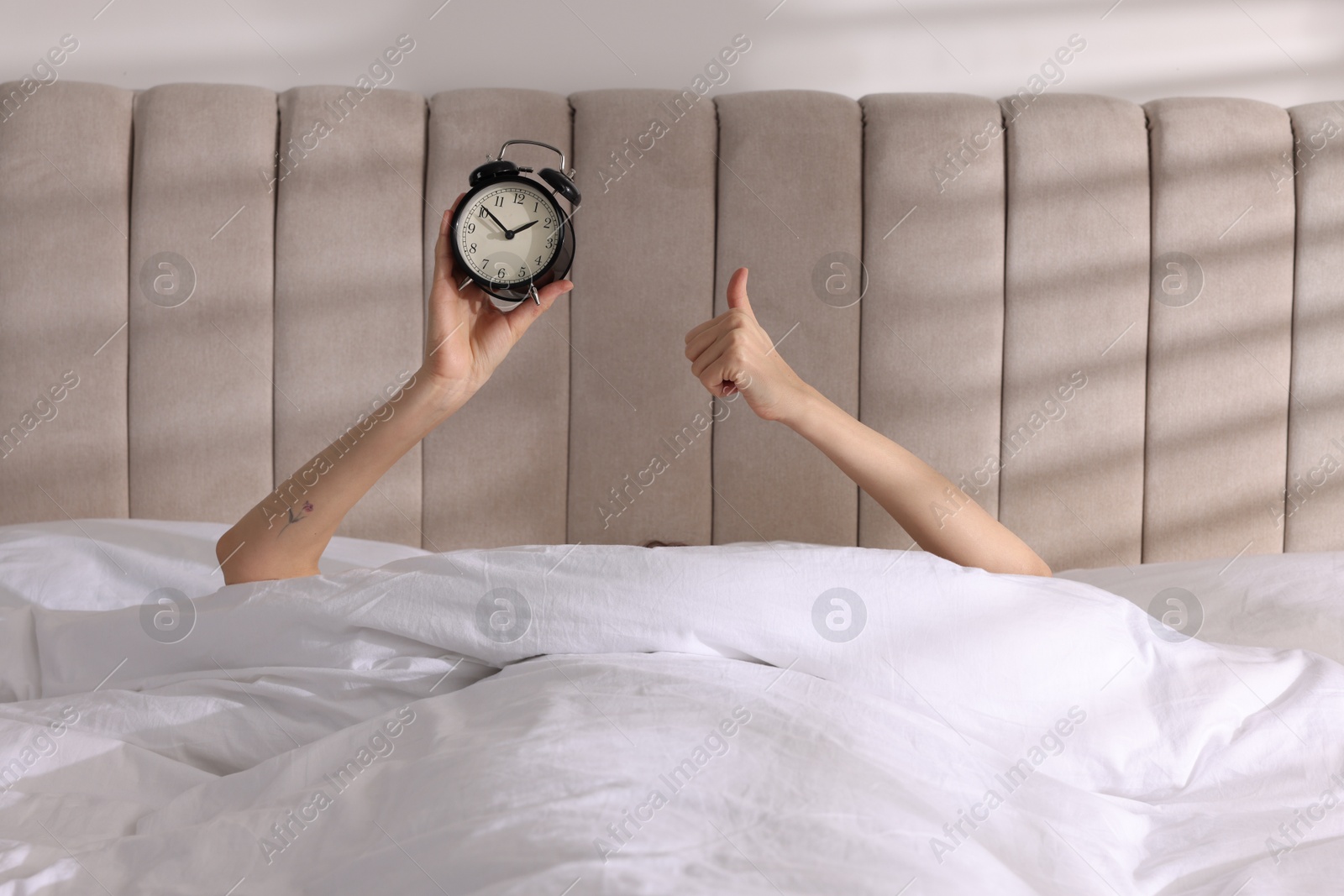 Photo of Woman with alarm clock showing thumbs up in bed at home, closeup. Good morning