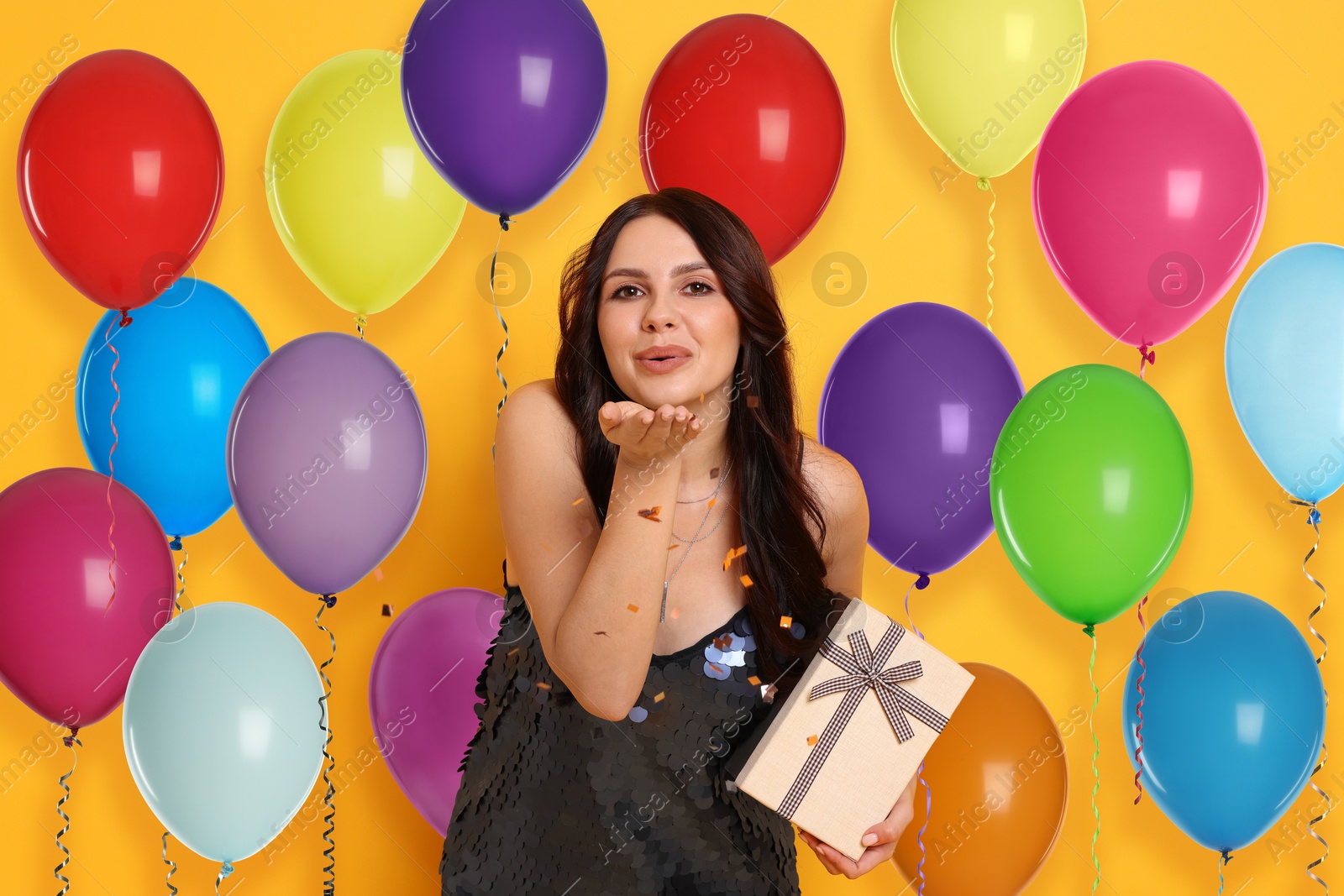 Image of Beautiful woman with gift box blowing confetti off hand on orange background. Surprise party with bright balloons