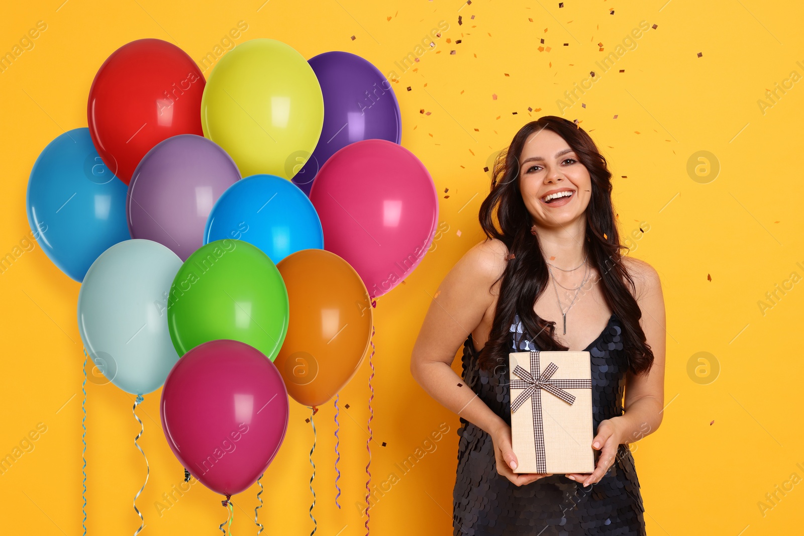 Image of Happy woman with gift box near bright balloons under falling confetti on orange background. Surprise party