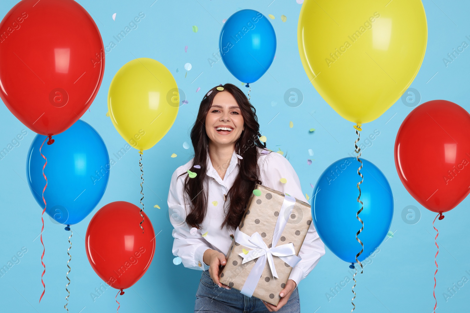 Image of Happy woman with gift box under falling confetti surrounded by bright balloons on light blue background. Surprise party