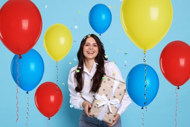 Image of Happy woman with gift box under falling confetti surrounded by bright balloons on light blue background. Surprise party