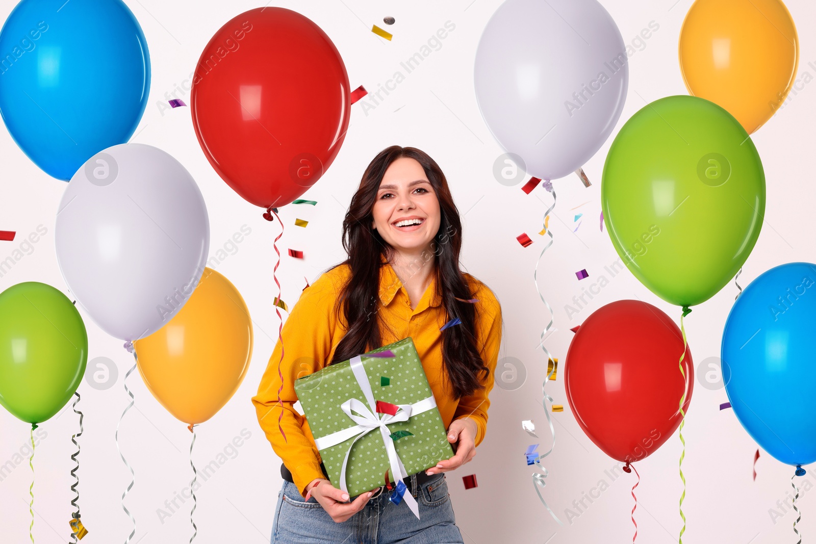 Image of Happy woman with gift box and falling confetti surrounded by bright balloons on white background. Surprise party