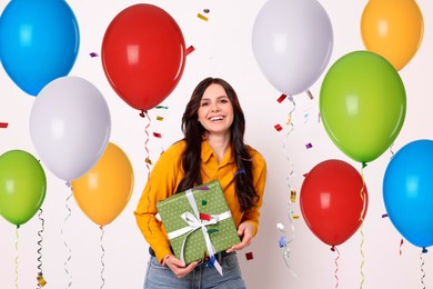 Image of Happy woman with gift box and falling confetti surrounded by bright balloons on white background. Surprise party