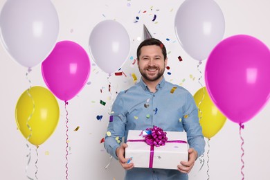 Image of Happy man with gift box under falling confetti surrounded by bright balloons on white background. Surprise party