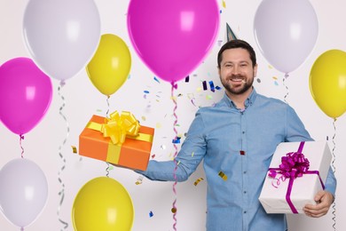 Image of Happy man with gift boxes under falling confetti surrounded by bright balloons on white background. Surprise party