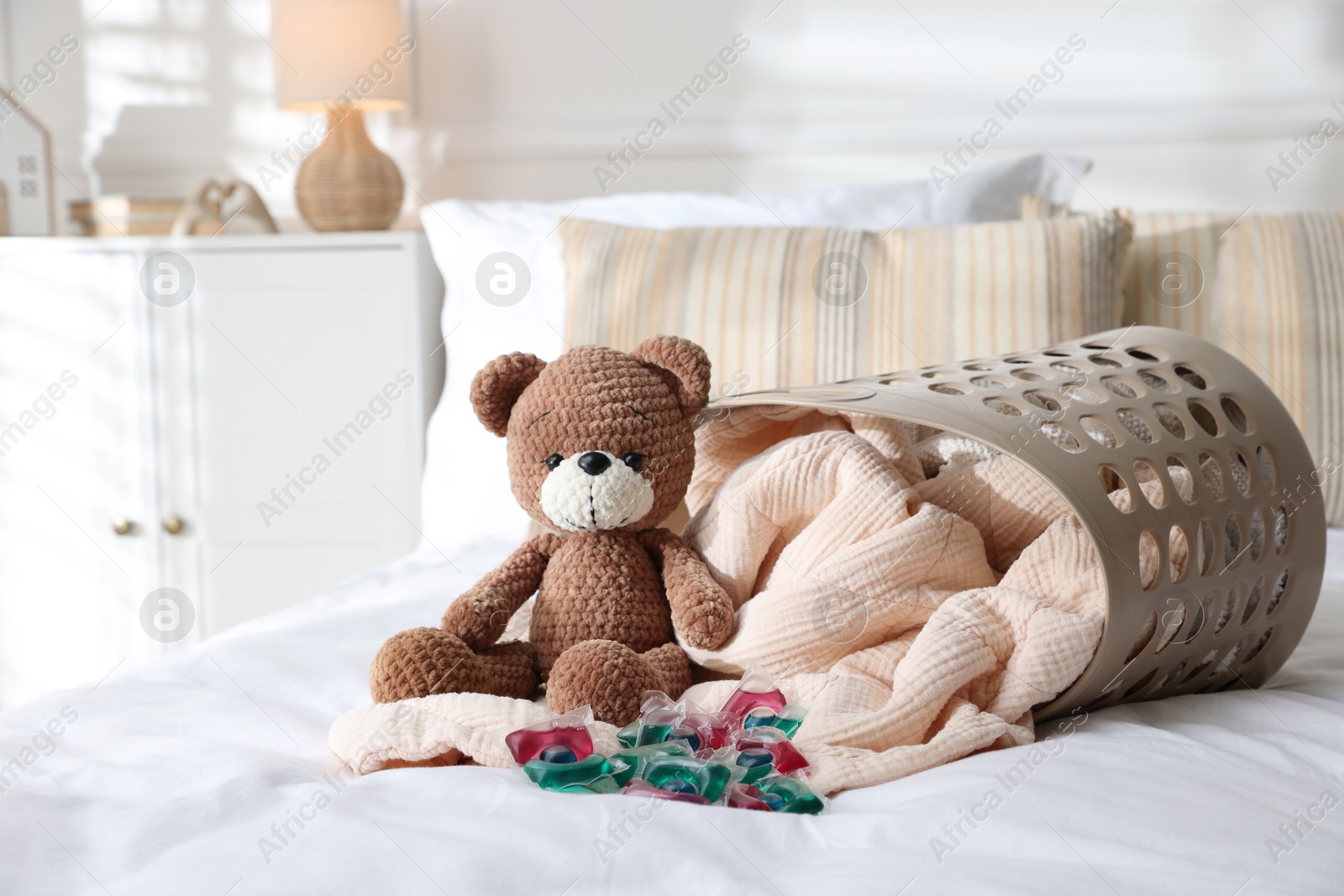 Photo of Laundry detergent capsules, teddy bear and basket with clean linens on bed indoors