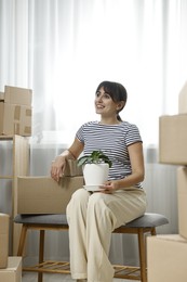 Photo of Moving day. Happy woman with houseplant and cardboard boxes in her new home