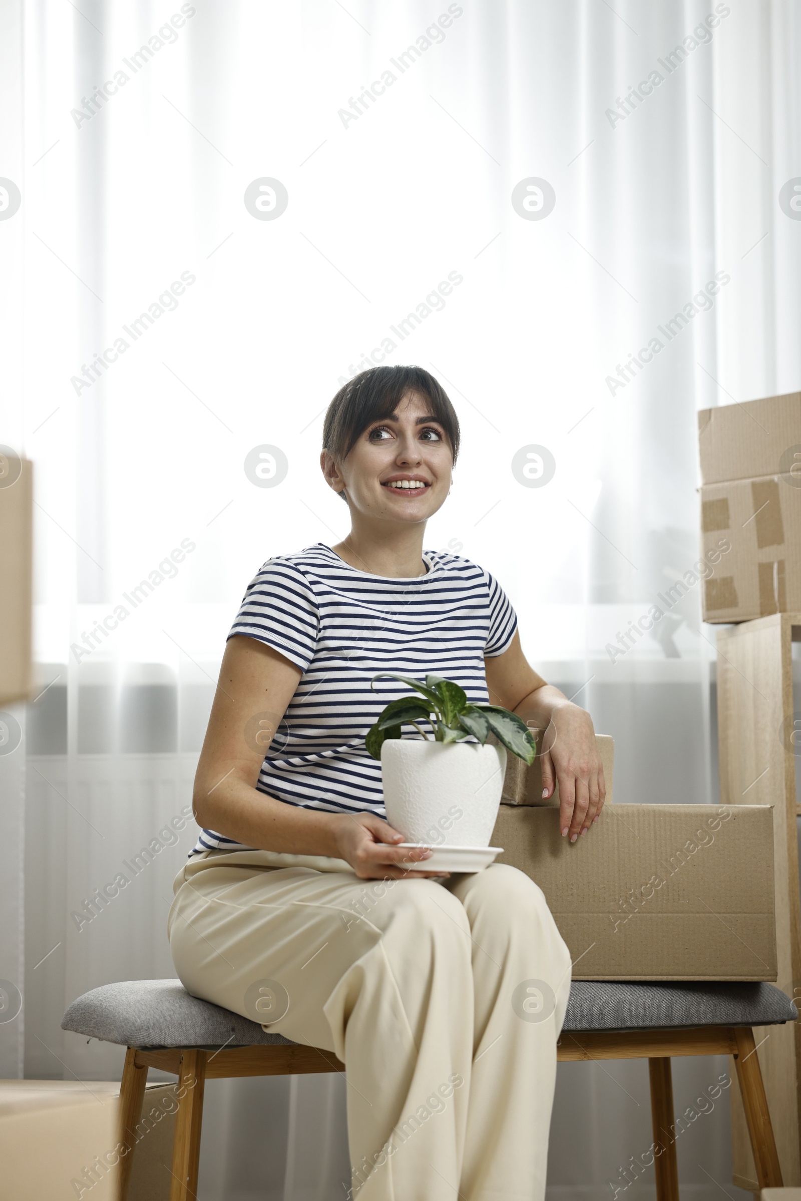 Photo of Moving day. Happy woman with houseplant and cardboard boxes in her new home