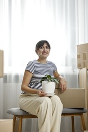 Photo of Moving day. Happy woman with houseplant and cardboard boxes in her new home