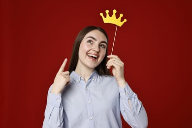 Photo of Happy woman holding stick with paper crown and pointing upwards on dark red background