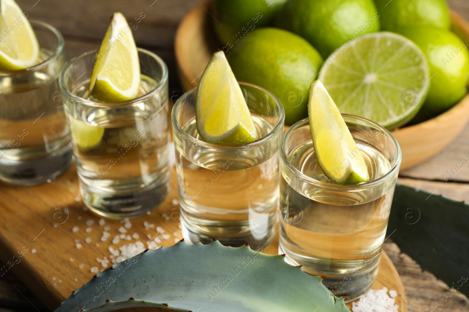 Photo of Tequila shots with lime slices, salt and agave leaves on wooden table, closeup