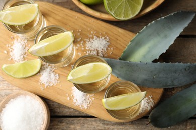 Photo of Tequila shots with lime slices, salt and agave leaves on wooden table, flat lay