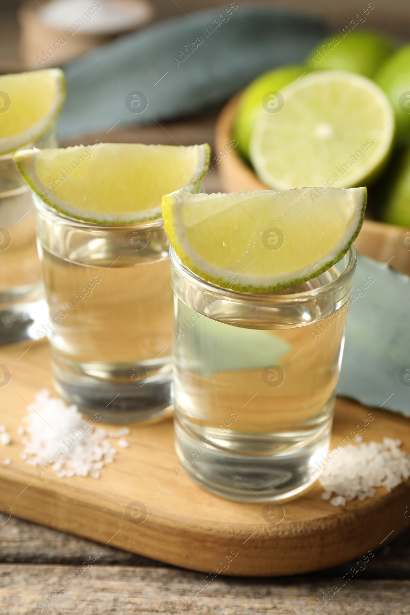 Photo of Tequila shots with lime slices, salt and agave leaves on wooden table, closeup