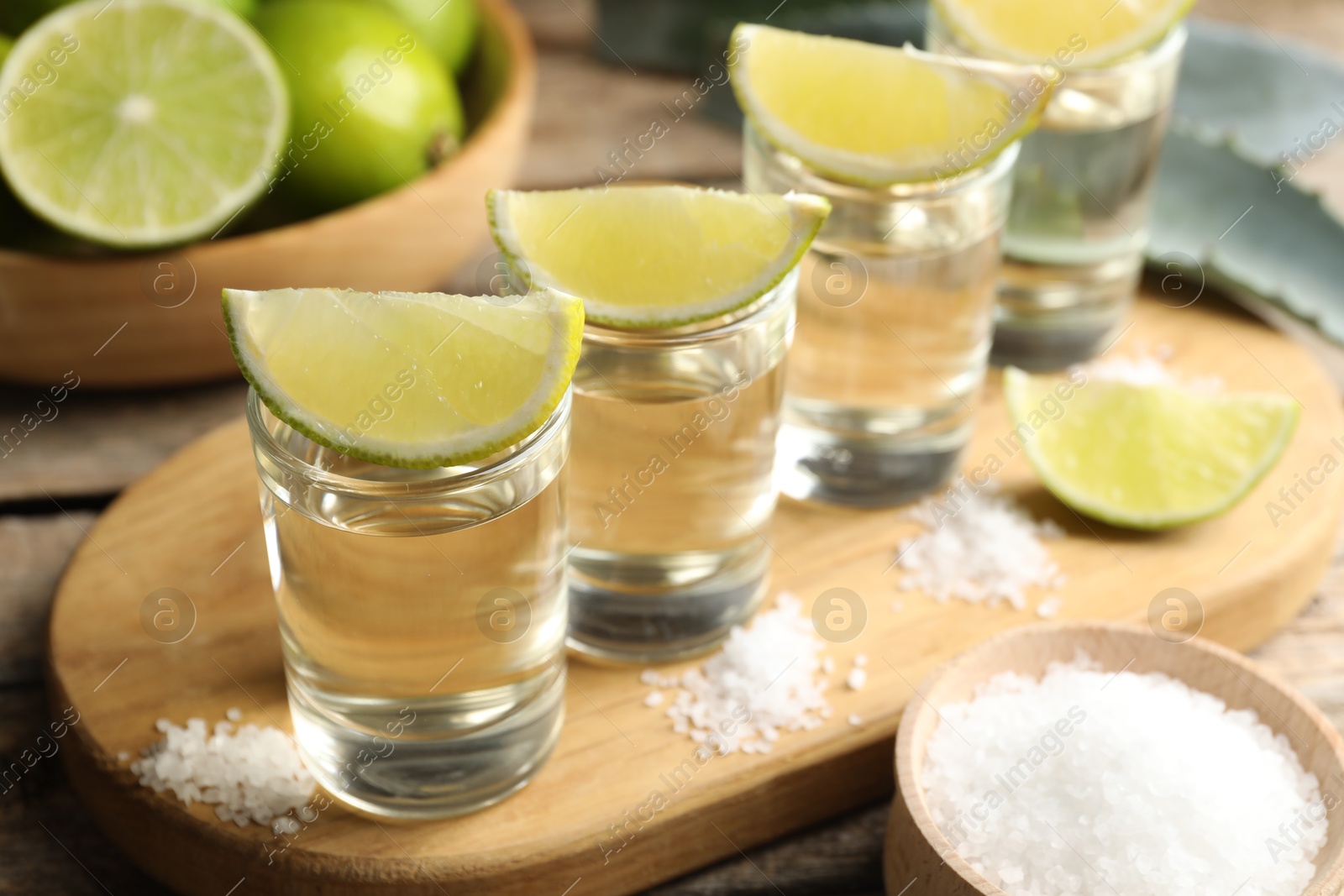 Photo of Tequila shots with lime slices, salt and agave leaves on wooden table, closeup