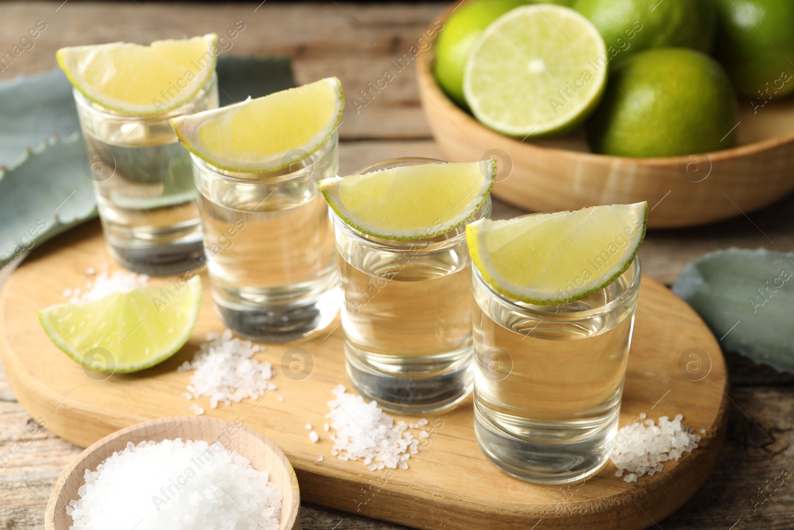 Photo of Tequila shots with lime slices, salt and agave leaves on wooden table, closeup