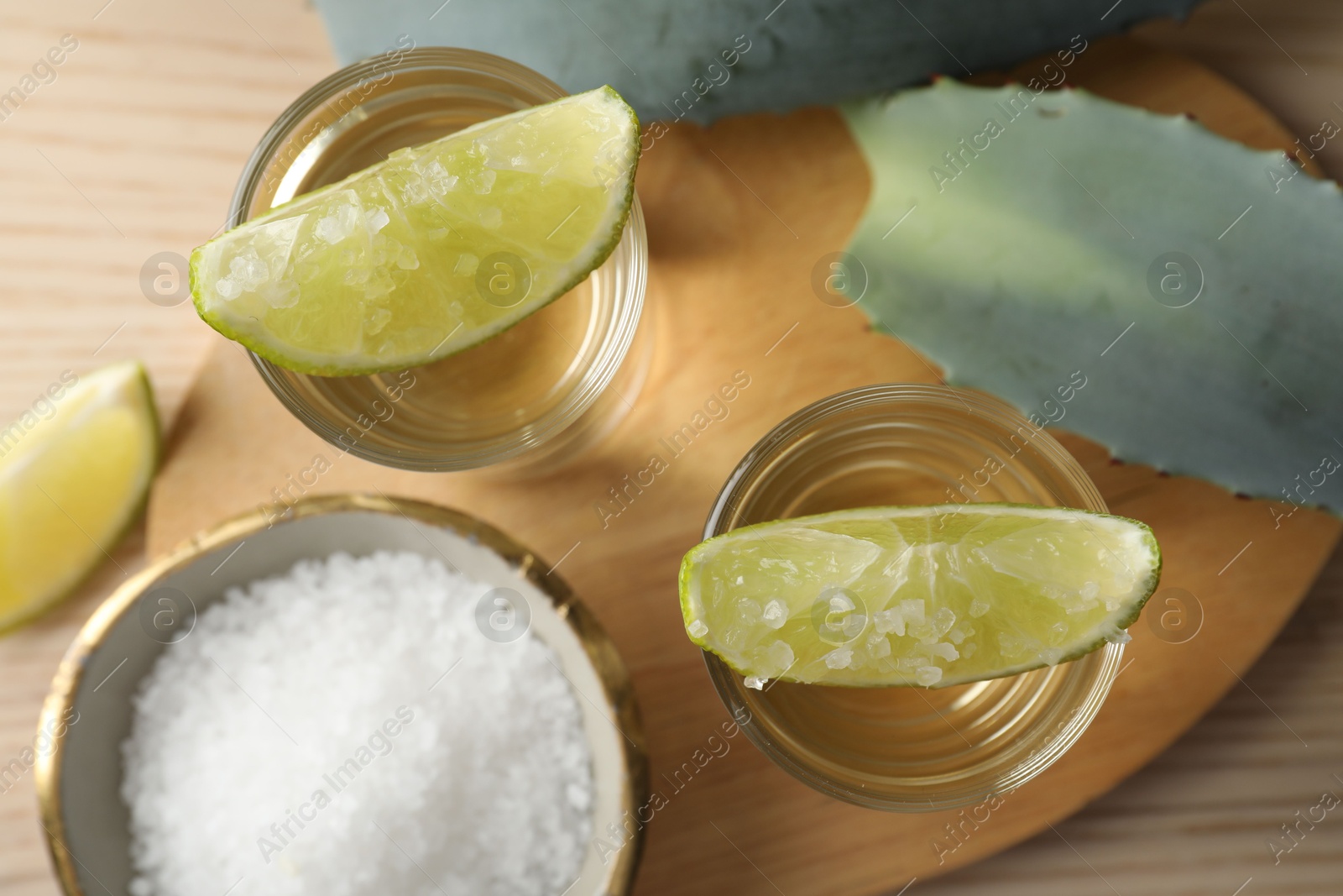 Photo of Tequila shots with lime slices, salt and agave leaves on wooden table, flat lay