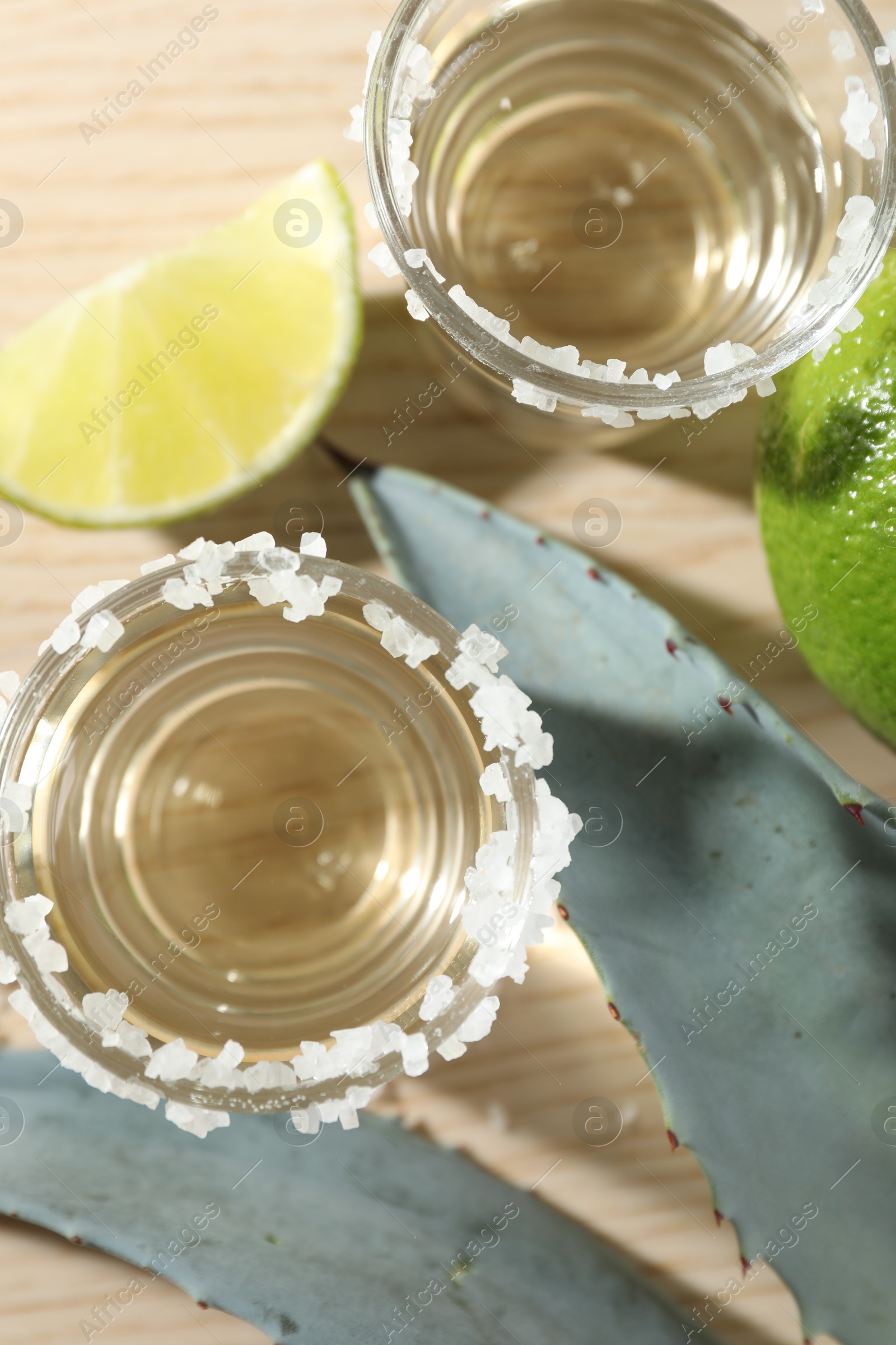 Photo of Tequila shots with salt, limes and agave leaves on wooden table, flat lay