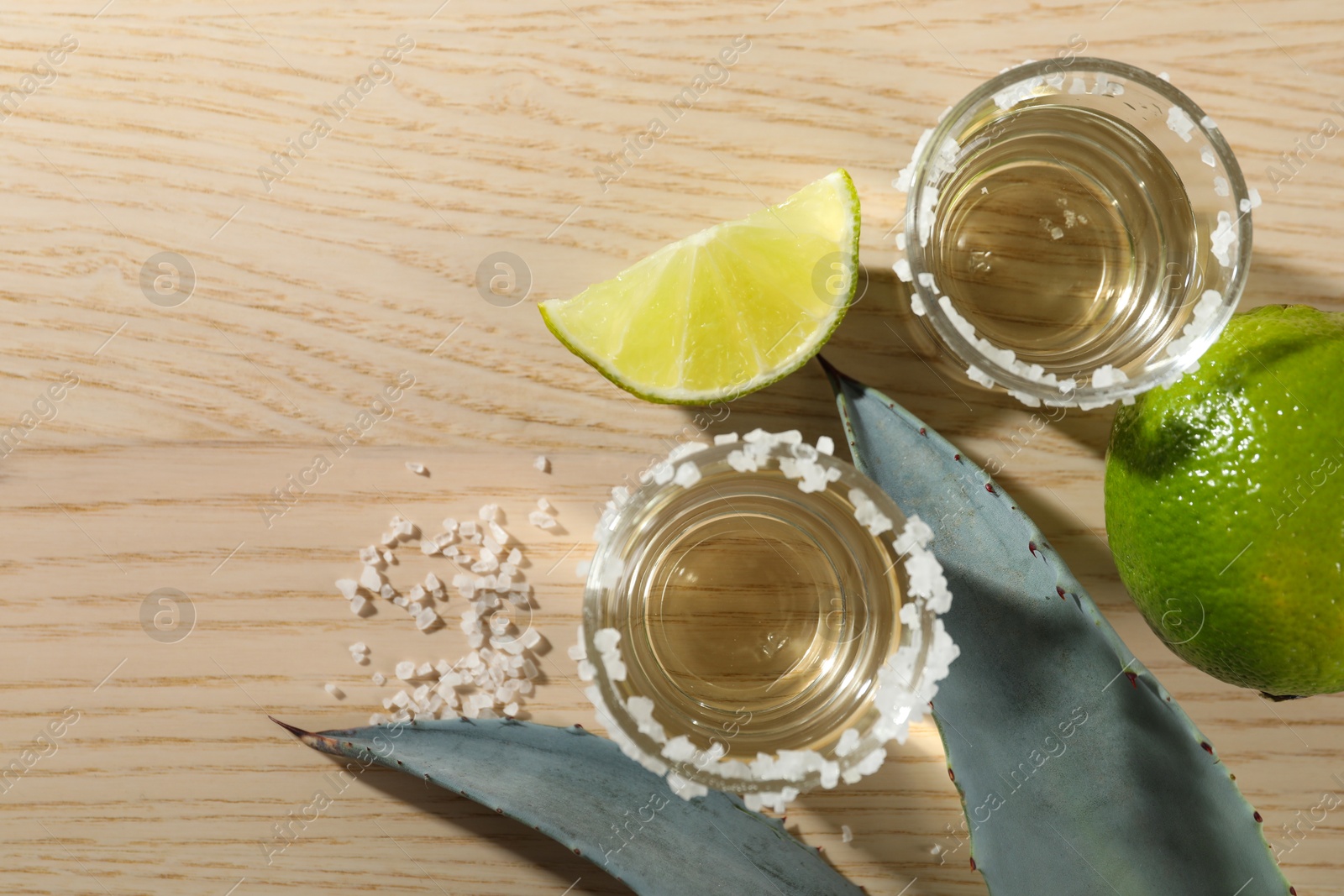 Photo of Tequila shots with salt, limes and agave leaves on wooden table, flat lay. Space for text