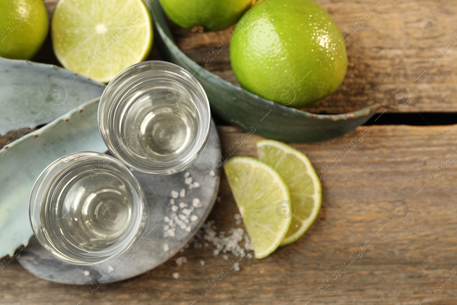 Photo of Tequila shots with salt, limes and agave leaves on wooden table, flat lay