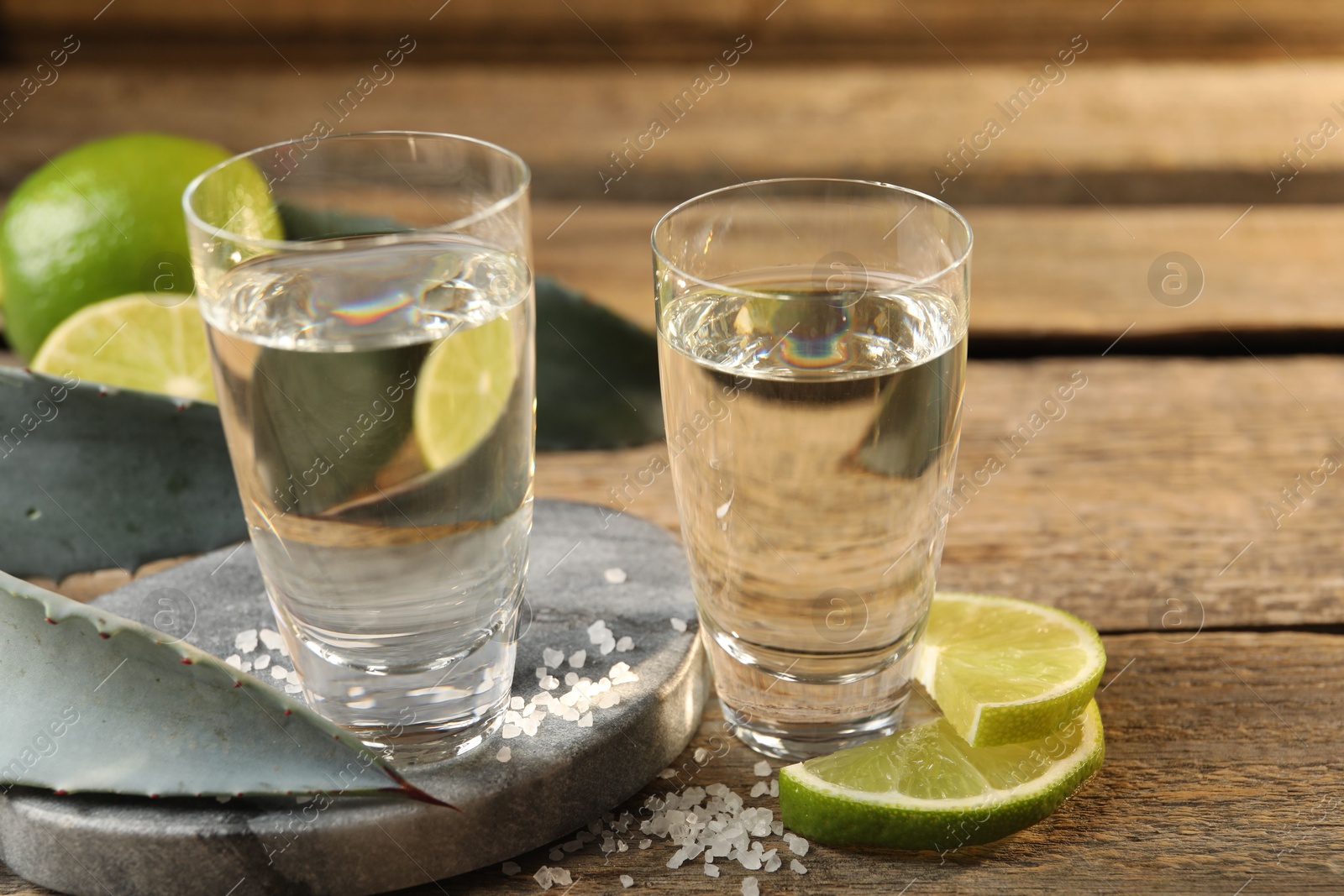 Photo of Tequila shots with salt, limes and agave leaves on wooden table, closeup