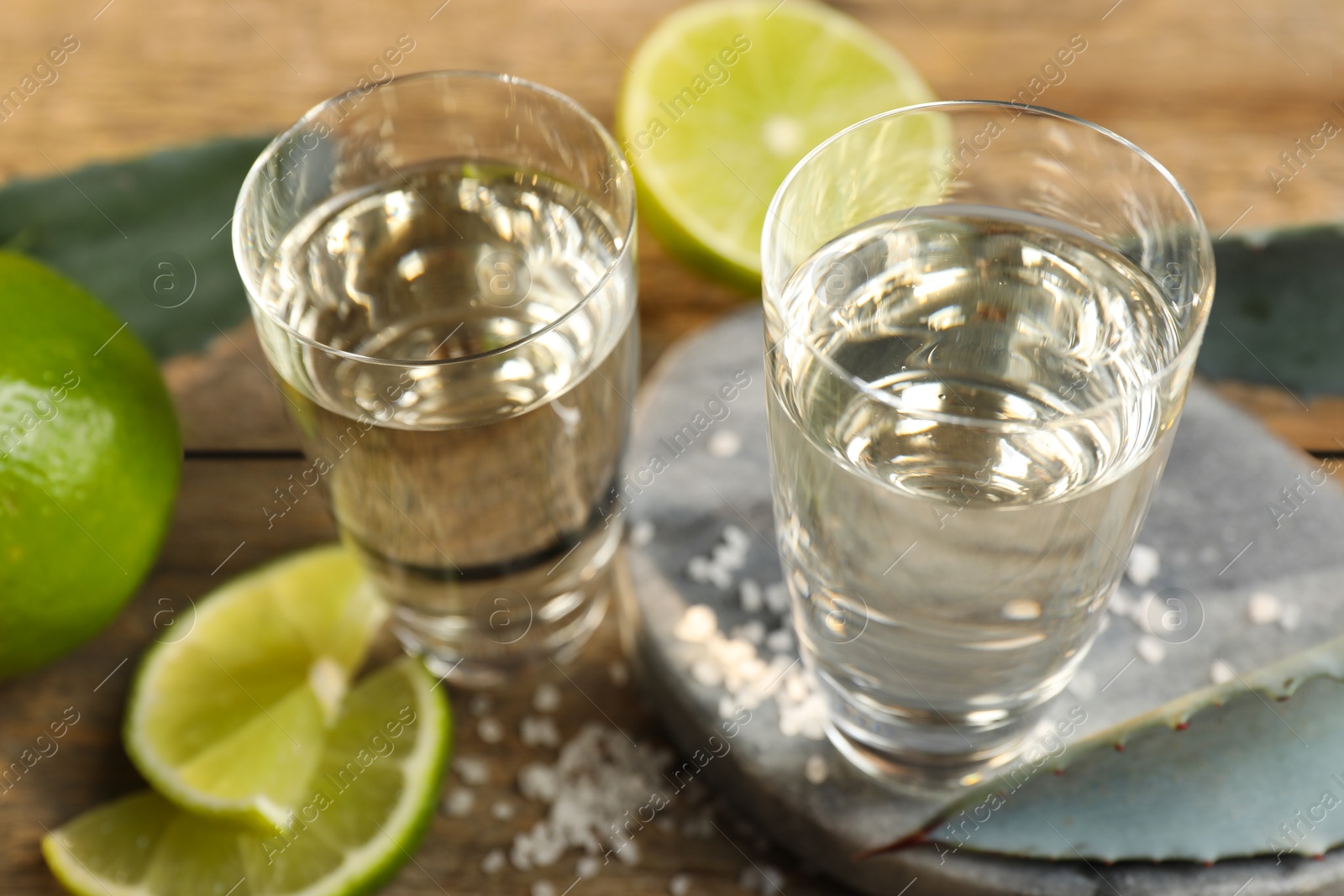 Photo of Tequila shots with salt, limes and agave leaves on wooden table, closeup