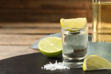 Photo of Tequila shot with lime slices, salt and agave leaves on wooden table, closeup. Space for text