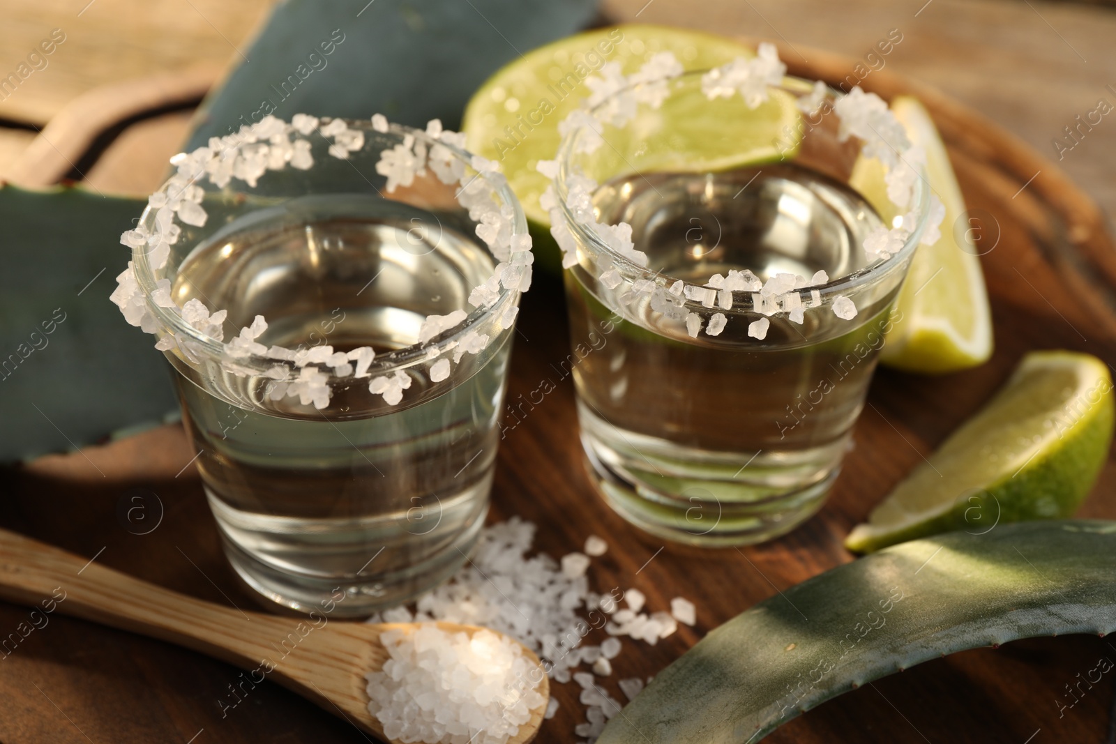 Photo of Tequila shots with salt, lime slices and agave leaves on wooden table, closeup