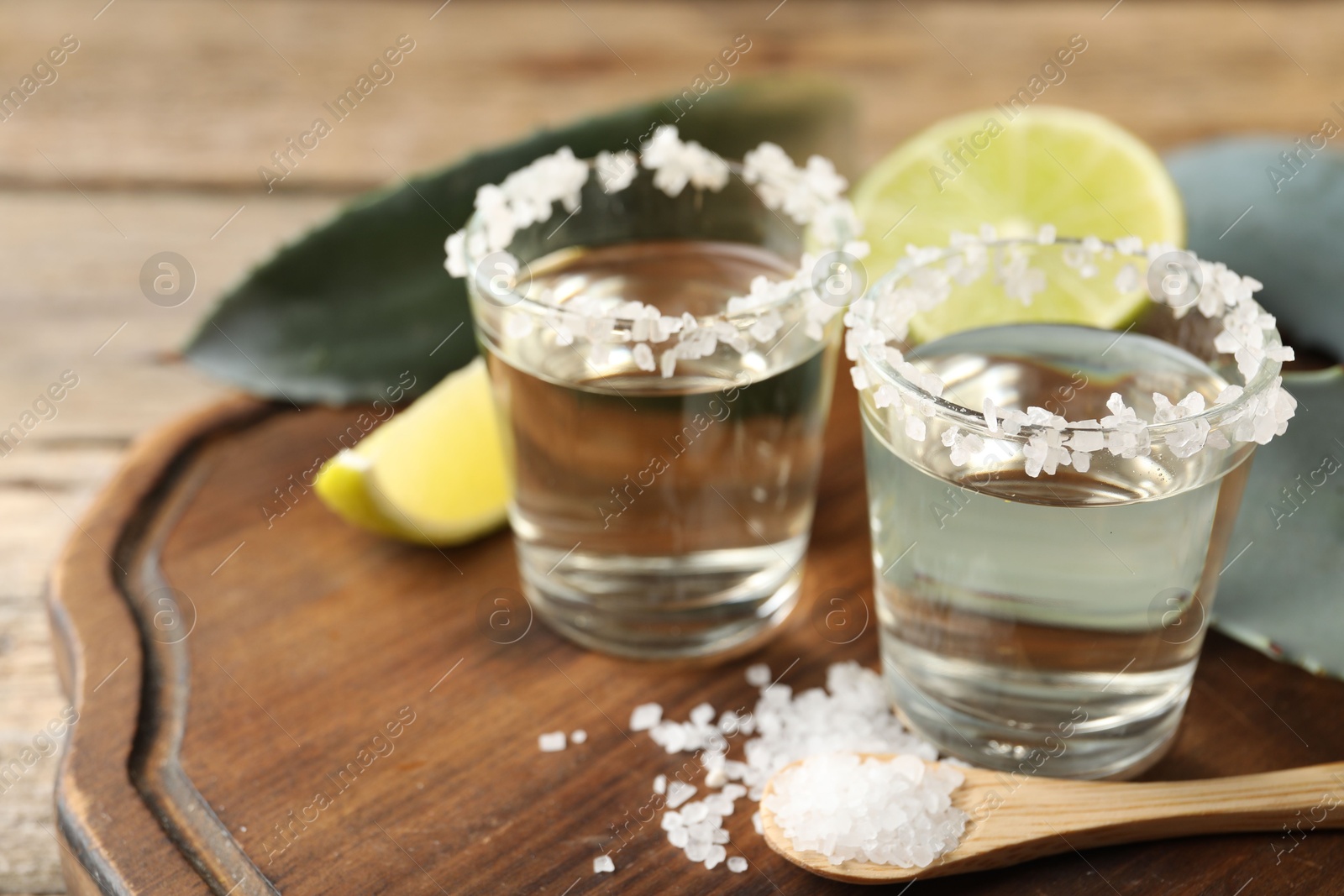 Photo of Tequila shots with salt, lime slices and agave leaves on wooden table, closeup