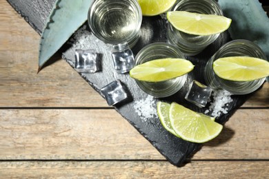 Photo of Tequila shots with lime slices, salt, ice cubes and agave leaves on wooden table, flat lay. Space for text