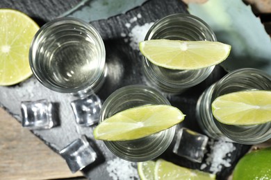 Photo of Tequila shots with lime slices, salt, ice cubes and agave leaf on wooden table, flat lay