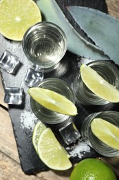 Photo of Tequila shots with lime slices, salt, ice cubes and agave leaves on wooden table, flat lay