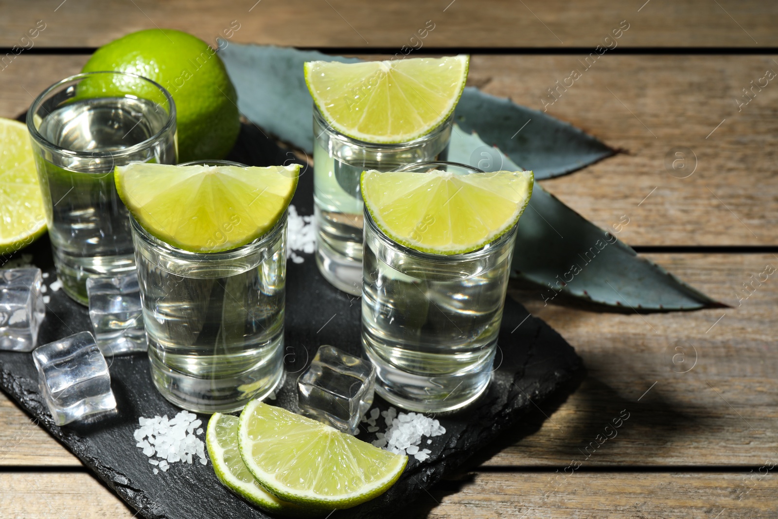 Photo of Tequila shots with lime slices, salt, ice cubes and agave leaves on wooden table, closeup