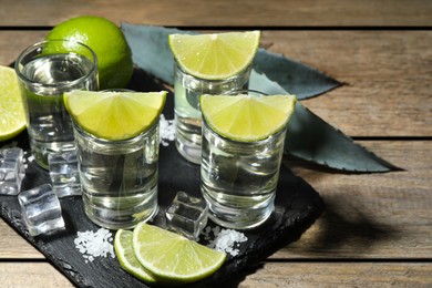 Tequila shots with lime slices, salt, ice cubes and agave leaves on wooden table, closeup