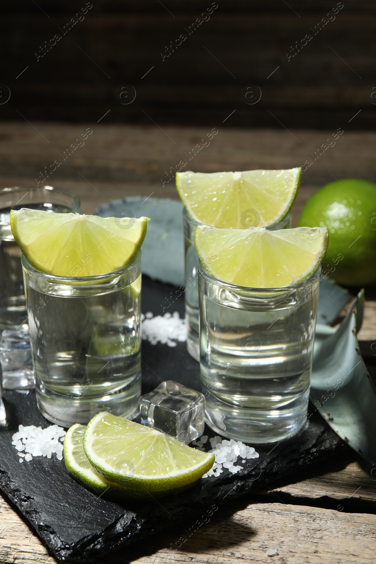 Photo of Tequila shots with lime slices, salt, ice cubes and agave leaf on wooden table, closeup