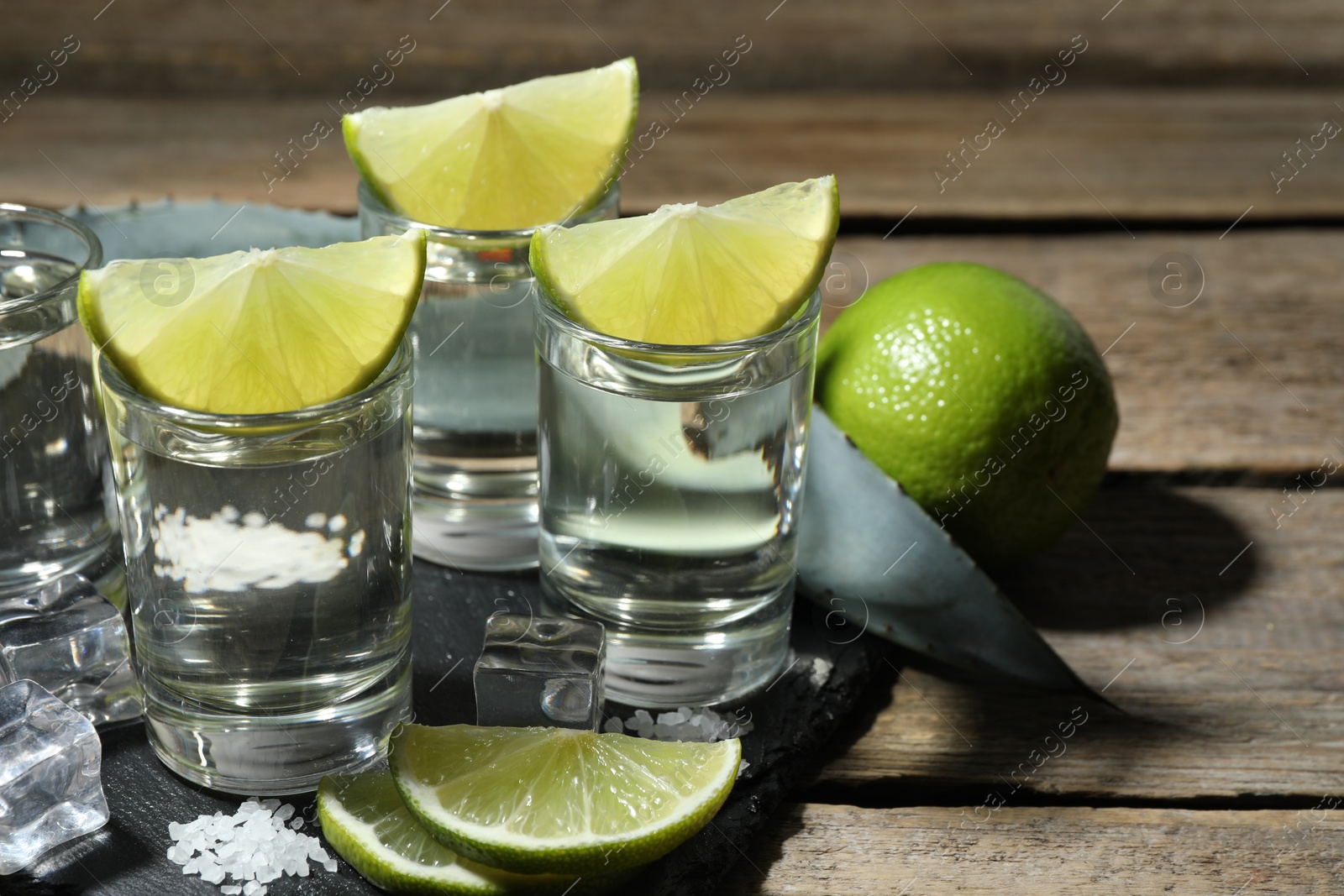 Photo of Tequila shots with lime slices, salt, ice cubes and agave leaf on wooden table, closeup