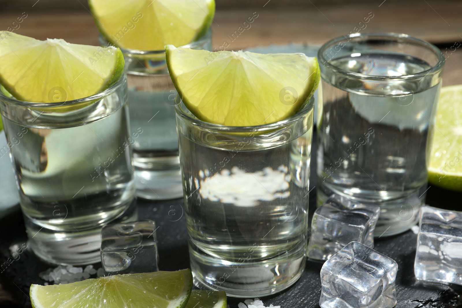 Photo of Tequila shots with lime slices, salt, ice cubes and agave leaf on table, closeup