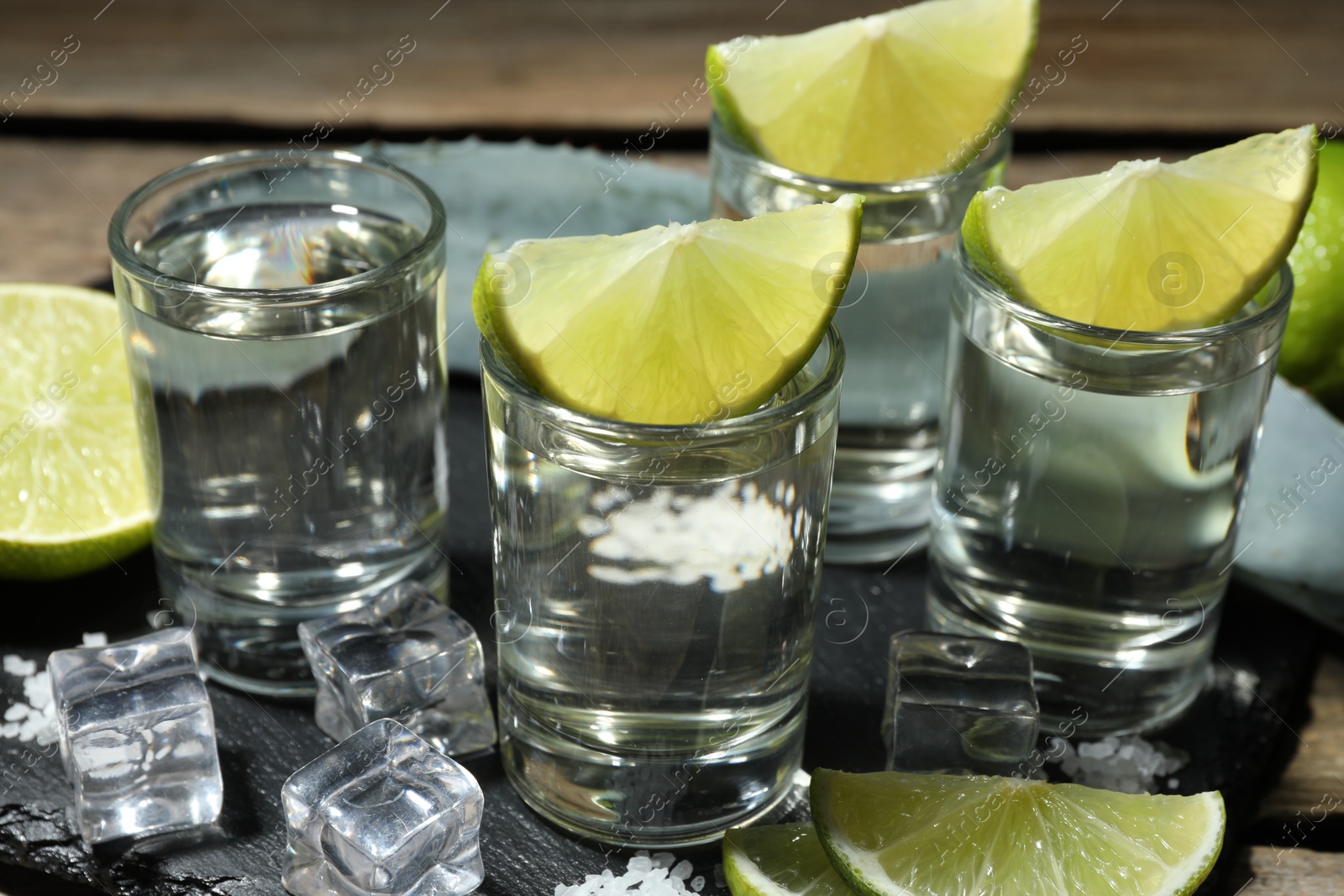 Photo of Tequila shots with lime slices, salt, ice cubes and agave leaf on table, closeup