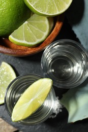 Photo of Tequila shots with lime slices, salt and agave leaves on table, flat lay